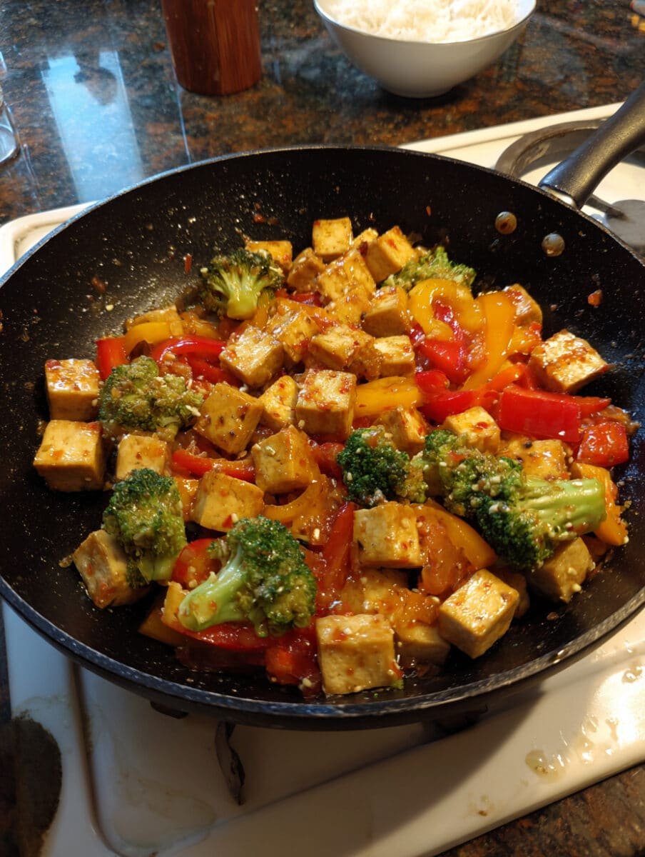 A bowl of sweet chili tofu stir-fry with colorful veggies coated in a glossy chili sauce, served as a quick vegan dinner.